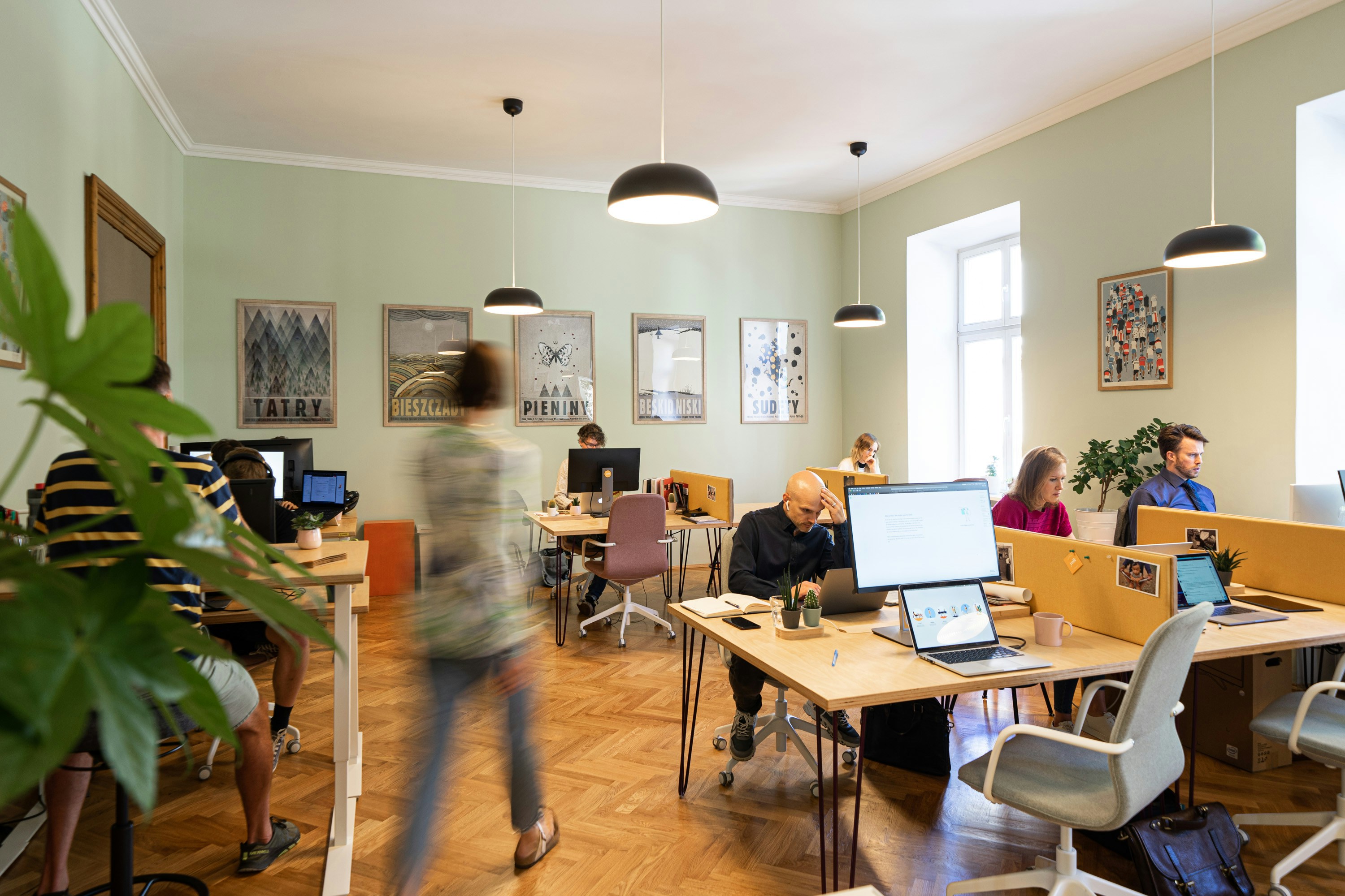 Image shows a busy office - with people working at their desks and facing computer screens. There is a person walking through the office space