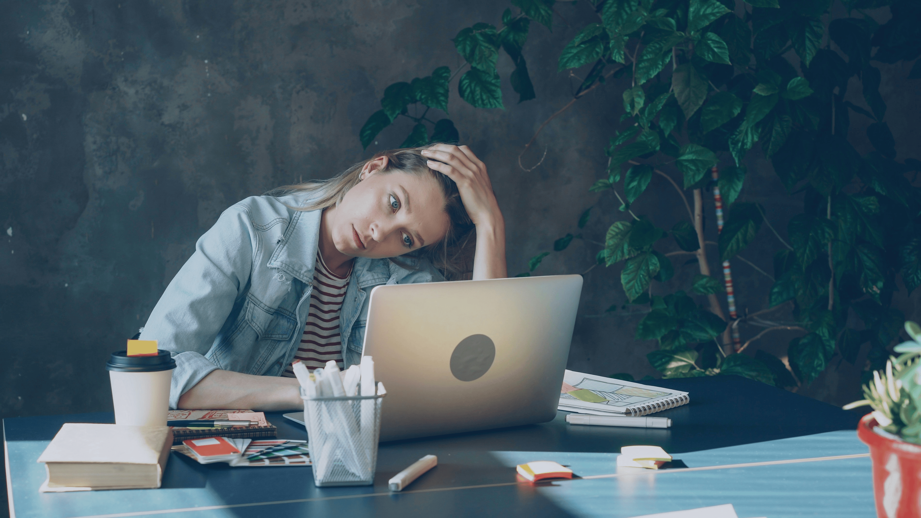 Image shows a woman at work, looking at her laptop screen, with her hand on her head. She is deep in thought.